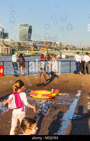 London, UK, 19. April 2018. Ein Entertainer am Südufer der Ufer der Themse in Bankside bläst Seifenblasen Kinder gegen eine Skyline Hintergrund von Ikonischen moderne Wolkenkratzer Gebäude in der Stadt London zu unterhalten. Die Sonnige schönem Wetter auf den wärmsten April Tag seit Jahrzehnten brachten gutmütige Massen, die Sonne zu genießen. Credit: Graham Prentice/Alamy Leben Nachrichten. Stockfoto