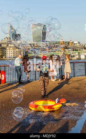 London, UK, 19. April 2018. Ein Entertainer am Südufer der Ufer der Themse in Bankside bläst Seifenblasen Kinder gegen eine Skyline Hintergrund von Ikonischen moderne Wolkenkratzer Gebäude in der Stadt London zu unterhalten. Die Sonnige schönem Wetter auf den wärmsten April Tag seit Jahrzehnten brachten gutmütige Massen, die Sonne zu genießen. Credit: Graham Prentice/Alamy Leben Nachrichten. Stockfoto