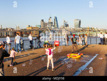 London, UK, 19. April 2018. Ein Entertainer am Südufer der Ufer der Themse in Bankside bläst Seifenblasen Kinder gegen eine Skyline Hintergrund von Ikonischen moderne Wolkenkratzer Gebäude in der Stadt London zu unterhalten. Die Sonnige schönem Wetter auf den wärmsten April Tag seit Jahrzehnten brachten gutmütige Massen, die Sonne zu genießen. Credit: Graham Prentice/Alamy Leben Nachrichten. Stockfoto