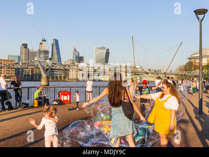 London, UK, 19. April 2018. Ein Entertainer am Südufer der Ufer der Themse in Bankside bläst Seifenblasen Kinder gegen eine Skyline Hintergrund von Ikonischen moderne Wolkenkratzer Gebäude in der Stadt London zu unterhalten. Die Sonnige schönem Wetter auf den wärmsten April Tag seit Jahrzehnten brachten gutmütige Massen, die Sonne zu genießen. Credit: Graham Prentice/Alamy Leben Nachrichten. Stockfoto