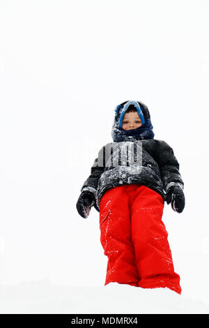 Auf der Suche auf ein Junge (5 Jahre alt) Outdoor Winter gekleidet. Er ist auf einem Haufen Schnee stand mit einem fast weißen Himmel hinter sich. Stockfoto