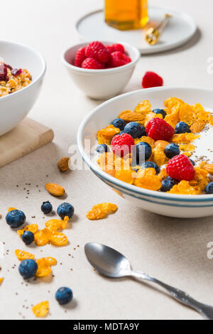 Gesundes Frühstück. Corn Flakes mit Joghurt, Mandeln, Blaubeeren, Himbeeren und Chia Samen. Mit Ahornsirup und Saft auf beiger Stoff serviert. Stockfoto