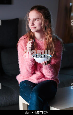 Blonde junge lächelnde Mädchen genießen Essen frische Blaubeeren Holding kleine Schüssel voller Früchte. Junges Mädchen trägt rosa Pullover und Jeans auf ta Sitzung Stockfoto