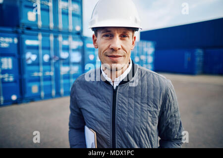 Lächelnd Ingenieur trägt einen Schutzhelm allein stehend auf einem großen kommerziellen Versand Dock eine Zwischenablage Stockfoto
