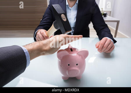 Der Geschäftsmann Hand anhalten sein Kollege vom Brechen Sparschwein mit Hammer im Büro Stockfoto