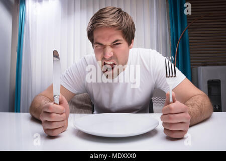 Wütend Mann mit Messer und Gabel mit leeren Teller am Tisch Stockfoto