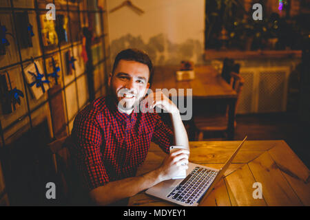 Eine junge Stattlichen kaukasischen Mann mit Bart und toothy Lächeln in rot kariertem Hemd arbeitet hinter Laptop an den hölzernen Tisch sitzen. Verwendet hält Mobile Stockfoto