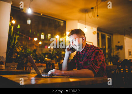 Junge stattlichen kaukasischen Mann mit Bart und toothy Lächeln im roten Hemd arbeitet hinter Laptop, Hände auf der Tastatur an den hölzernen Tisch sitzen. Verwendet, um die Anrufe auf Mo Stockfoto