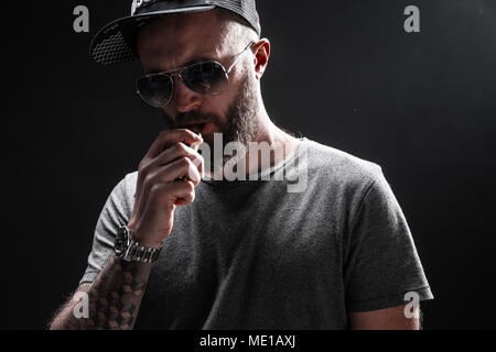 Der bärtige Mann gekleidet in einem grauen T-Shirt, Sonnenbrille und Baseballmütze vaping Stockfoto