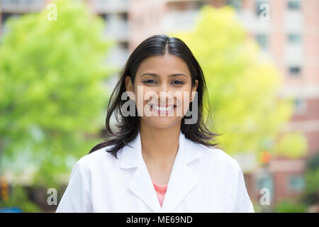 Closeup Portrait von zuversichtlich, lächelnd weibliche Ärztin im weißen Kittel, isolierten Hintergrund des unscharfen Bäume und Gebäude Stockfoto