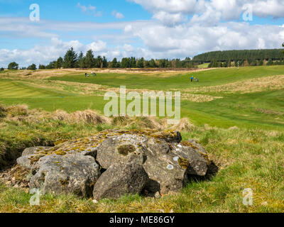 West Linton, Scottish Borders, Schottland, Vereinigtes Königreich, 21. April 2018. UK Wetter: Warme Frühlingssonne bei West Linton Golfplatz mit Golfspieler, die über die Fahrrinne. Bronzezeit cist im Vordergrund. Stockfoto