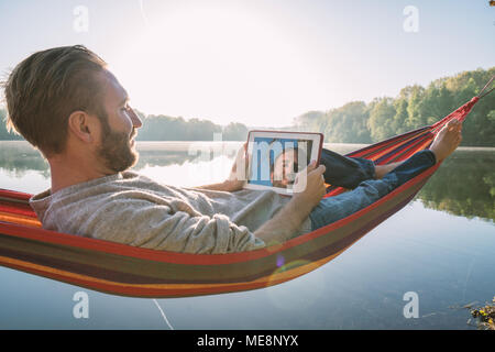 Fröhlicher junger Mann entspannt auf Hängematte mit digitalen Tablet am See am Morgen, schönen Sonnenlicht. Die Menschen reisen Technik Entspannung Konzept Stockfoto