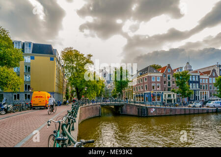 Eine niederländische Frau reitet ihr Fahrrad über eine Brücke über einen Kanal an einem bewölkten Herbst Tag in Amsterdam, Niederlande Stockfoto