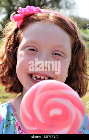 Eine rothaarige Behaarte Mädchen in ein schickes blaues Kleid mit einem großen, rosa und weißen Lollipop Stockfoto