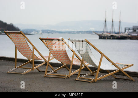 Liegestühle links am Kai in grauen und kalten Wetter. Mit einem Segelschiff. als Symbol für die Sehnsucht nach Sommer und Reisen. Träumen von Reisen und vacat Stockfoto