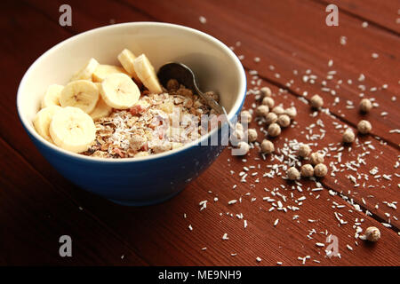 Gesundes Frühstück. Müsli Schale mit Banane, Rosinen und Nüssen. Und ein Glas Milch auf dem Holz- Hintergrund. Nahaufnahme Foto. Sweet vegane Gerichte. Morgen Stockfoto