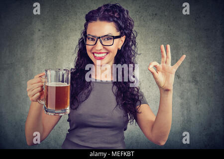 Glückliche Frau in Brillen gerne posieren bei Camera holding Becher Bier, ok Zeichen Stockfoto