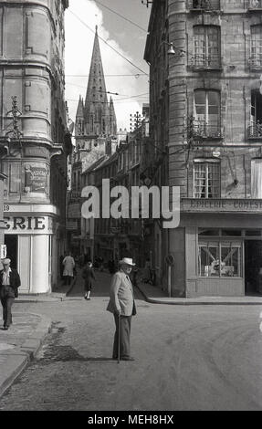 1950, historische, eine ältere gentlemam mit einem Spazierstock kreuzt eine Straße im Zentrum der Stadt in der Normandie Stadt Caen, Frankreich. Ein Turm der Abbaye Aux Hommes kann in der Ferne sehen. Stockfoto