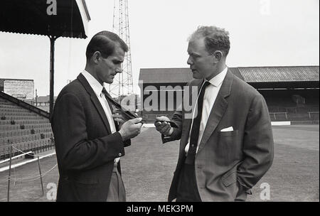 1964, Charlton Athletic FC, historisches Bild, auf dem Spielfeld im Valley zeigt ein Charlton-Fußballer einem lokalen Zeitungsreporter seine neue offizielle Krawatte des Fußballclubs. Zwischen 1964 und 1966 trugen die Charlton-Spieler ein neues Trikot, ein komplett weißes Trikot mit roten Schultern. Das Hand- und Schwertabzeichen erschien zuerst auf dem Fußballtrikot und auf der Clublie. Stockfoto