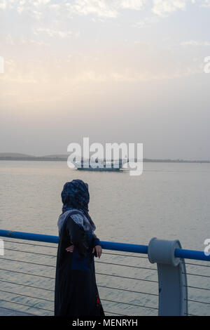 Eine asiatische Frau mit traditionellen arabischen Kleid (abbaya) und Schal, ein Boot in der Entfernung am offenen Meer, Abu Dhabi, VAE Stockfoto