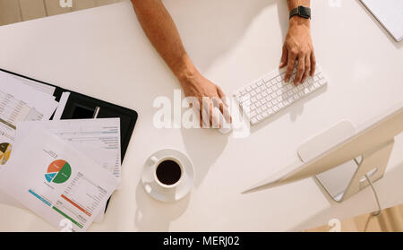 Blick von oben auf ein Mann bei der Arbeit am Computer mit Dokumenten und eine Tasse Kaffee auf den Tisch. Mit der drahtlosen Tastatur und Maus während der Arbeit am Computer Mann Stockfoto