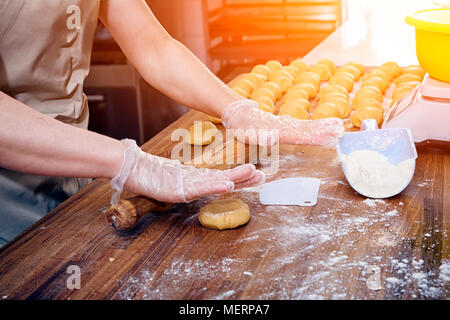Eine Nahaufnahme eines weiblichen Baker in Schutzhandschuhe zieht den Teig für das Kochen Brötchen auf einer verknoteten Tabelle mit einem Nudelholz Stockfoto