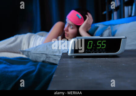 Bild der Frau mit Schlaflosigkeit mit rosa Bandagen für Augen liegend auf Bett neben Wecker Stockfoto