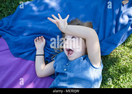 Vier Jahre alte blonde Mädchen liegt auf Handtücher in das grüne Gras Park mit Hand in lachend den Kopf mit lustigen Ausdruck Gesicht necken Stockfoto
