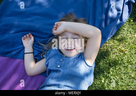 Vier Jahre alte blonde Mädchen liegt auf Handtücher in das grüne Gras Park mit Hand in lachend den Kopf mit geschlossenen Augen Stockfoto