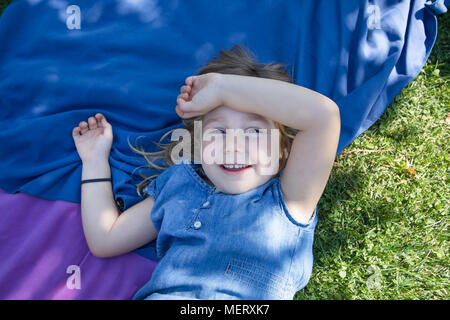 Vier Jahre alte blonde Mädchen liegt auf Handtücher in das grüne Gras der Park mit der Hand in den Kopf schauen und lächeln Stockfoto