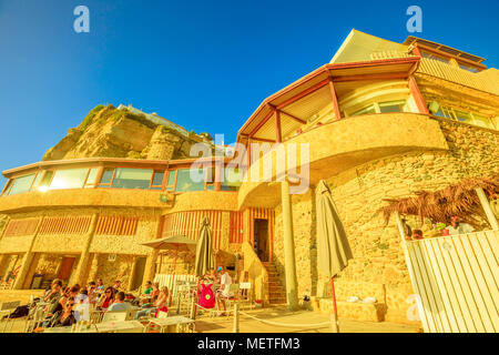 Azenhas do Mar, Portugal - August 5, 2017: Ansicht von unten auf das Restaurant und der hohen Klippe des ehemaligen Fischerdorf Azenhas do Mar in Colares an der Atlantikküste, vom Strand aus gesehen. Abendlicht. Stockfoto