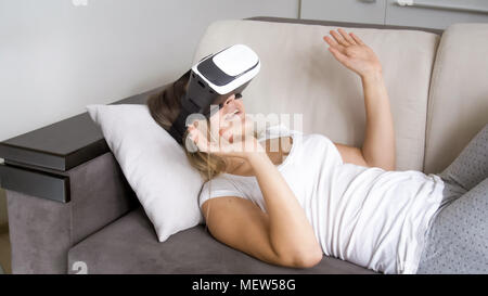 Glückliche junge Frau liegt auf einem Sofa und mit Virtual reality Headset Stockfoto