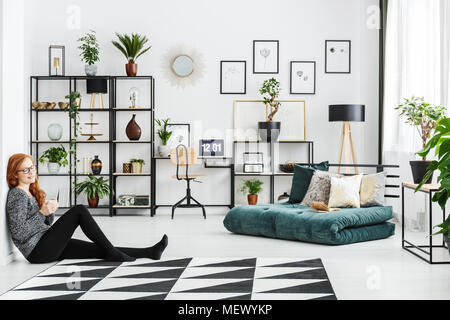 Junge Frau in Gläsern sitzen auf dem Boden und einen Becher in helles Wohnzimmer Interieur mit Laptop Stockfoto