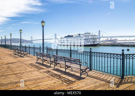 Bänke an historischen Pier 7 mit traditionellen Tretboot und die Oakland Bay Bridge im Hintergrund an einem sonnigen Tag mit blauen Himmel und Wolken, San Francisco Stockfoto