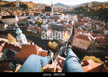 Antenne Birds Eye View junger Bergsteiger auf dem Dach sitzt auf der Kante von einem hohen Turm seine Füße hoch über der Altstadt von Cesky Krumlov baumelt. Stockfoto