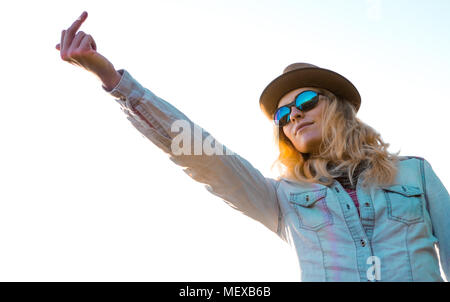 Portrait einer jungen kaukasischen Frau trägt einen stylischen Hut und Sonnenbrille stehen im Freien und mit einem Mittelfinger, Stockfoto