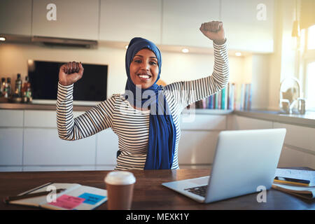 Junge muslimische Frauen Unternehmer trägt ein Kopftuch, das ihre Arme und feiern Ihren geschäftlichen Erfolg während der Arbeit auf einem Notebook in Ihrer Küche Stockfoto