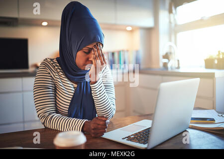 Die junge arabische weibliche Unternehmer Tragen eines hijab an ihrem Küchentisch sitzen, das online arbeitet mit einem Laptop betonte Stockfoto