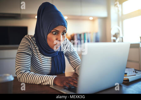 Junge arabische Frau das Tragen eines hijab schockiert schauen, während Sie online mit einem Laptop in Ihrer Küche zu Hause arbeiten Stockfoto