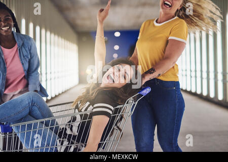 Lachende junge Frau in einem Warenkorb Sie einen Gang in die Stadt, die von zwei Freundinnen gedrückt wird Stockfoto