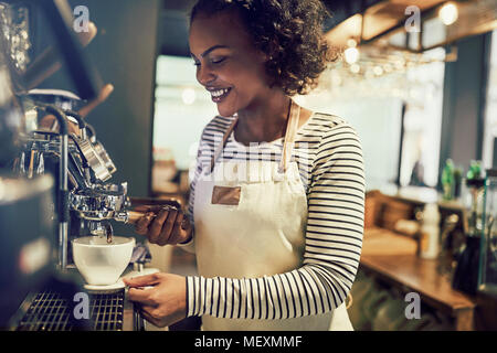 Lächelnden jungen afrikanischen barista Vorbereitung eine frische Tasse Kaffee, während Sie sich an einer Kaffeemaschine in einem angesagten Café stehend Stockfoto