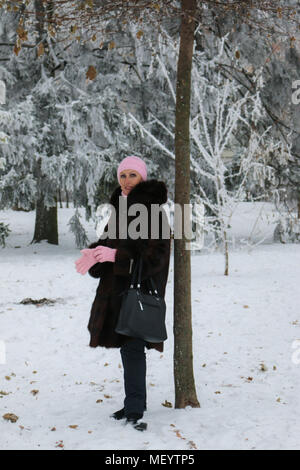 Lächelnde Dame in einem Pelzmantel und rosa Handschuhe im Winter Wald unter Schneefall. Stockfoto