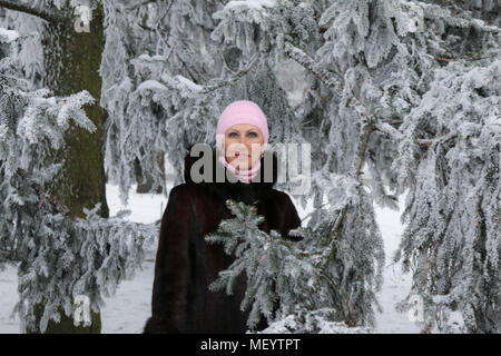 Schönen lächelnden Frau mittleren Alters in Pelzmantel und rosa Winter hat unter den Schnee - Bäume Stockfoto