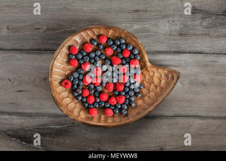 Table Top Aussicht auf eine gemischte Heidelbeeren und Himbeeren in Vintage geschnitzte hölzerne Platte auf grau Holz Schreibtisch platziert. Stockfoto