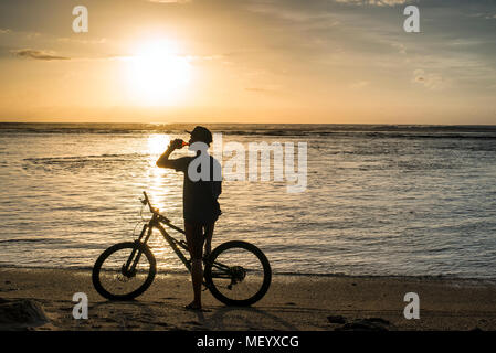 Ein Mann auf einem Mountainbike genießt ein Bier am Strand von Saint-Gilles auf der Insel Reunion im Indischen Ozean bei Sonnenuntergang. Stockfoto