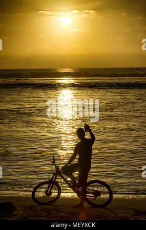 Ein Mann auf einem Mountainbike genießt ein Bier am Strand von Saint-Gilles auf der Insel Reunion im Indischen Ozean bei Sonnenuntergang. Stockfoto