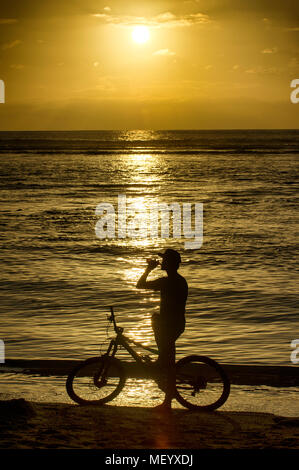 Ein Mann auf einem Mountainbike genießt ein Bier am Strand von Saint-Gilles auf der Insel Reunion im Indischen Ozean bei Sonnenuntergang. Stockfoto