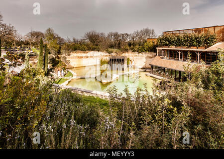 Der Saunagarten des Hotel Adler Thermae liegt in einem ehemaligen Steinbruch nahe Bagno Vignoni, Toskana, Italien; eine hölzerne Saunakabine steht auf Stelzen über einem grünlichen Quellbecken, das von freigelegten Felswänden eingerahmt ist, mit der Gartenterrasse des Hotelrestaurants rechts. Im Stil einer toskanischen Villa gestaltet, wurde das Resort in denselben Steinbruch gebaut und fügt sich in die Landschaft der Val d'Orcia ein, die seit 2004 UNESCO-Weltkulturerbe ist. Stockfoto