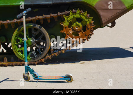 Kinder scooter vor einem Teil eines Caterpillar Track auf eine militärische Amphibienfahrzeug. Das Fahrzeug hat eine grüne camouflage Scheme. Stockfoto