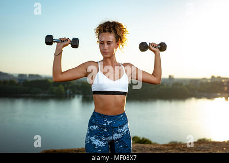 Junge fitness Frau tun Training mit Hanteln durch den Fluss in einem Sonnenuntergang. Stockfoto